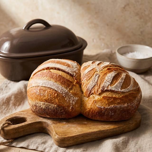 Zwei goldbraune, zusammengewachsene Brotlaibe mit knuspriger, bemehlter Kruste auf einem Holzbrett, im Hintergrund der Pampered Chef® Ofenmeister