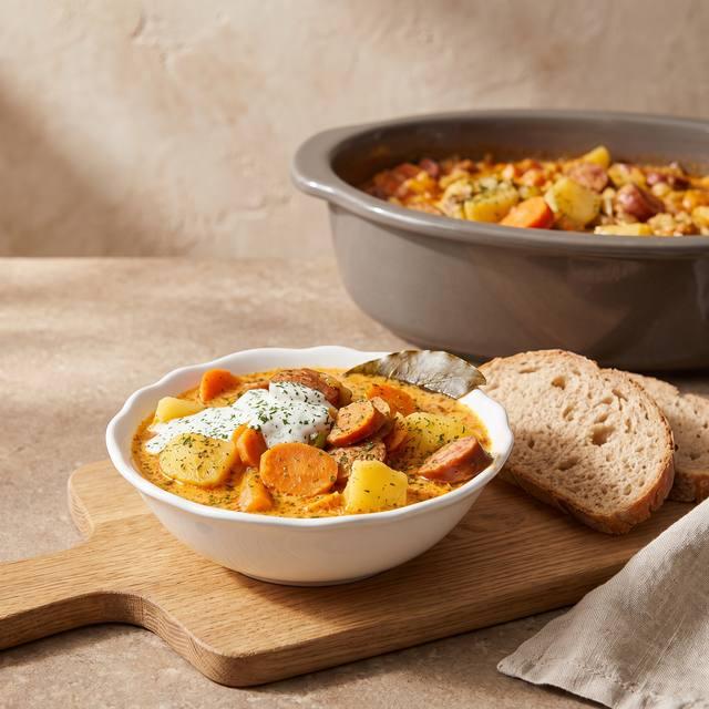 Krakauer-Eintopf mit Kartoffeln, Möhren und Kräuter-Sahne in einer weißen Schüssel, im Hintergrund der Pampered Chef® Ofenmeister