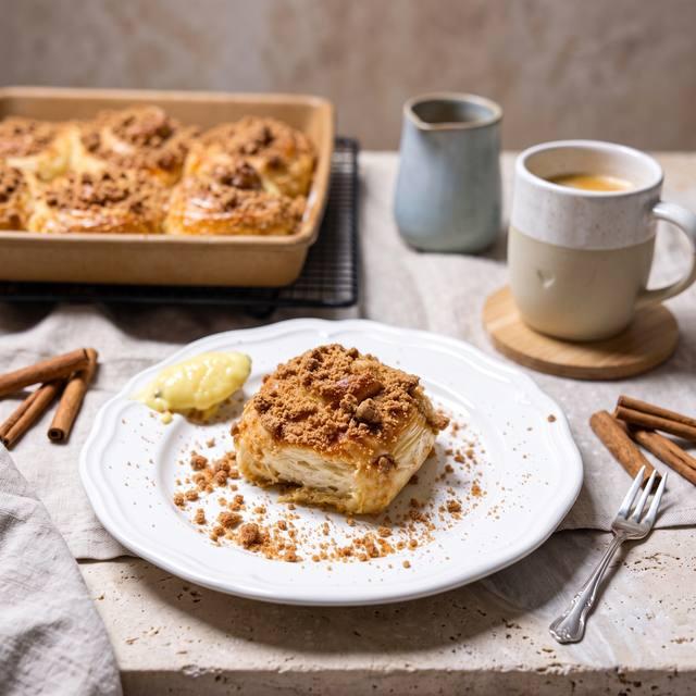 Goldbraune Spekulatius-Schnecke mit Frischkäse-Frosting und Spekulatiuskrümeln auf einem weißen Teller, im Hintergrund das Blech mit weiteren Schnecken – Pampered Chef® Rezept