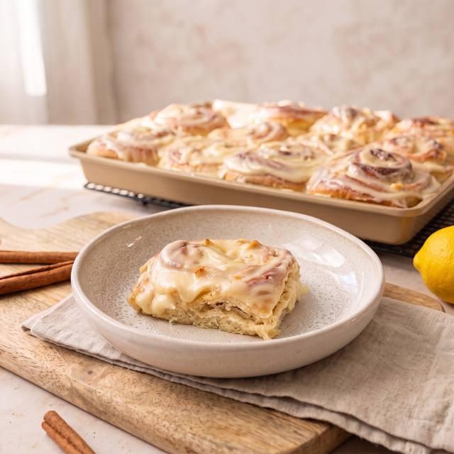 Vanille-Apfel-Zimtschnecke mit Frischkäse-Frosting auf einem Teller, im Hintergrund das volle Blech – Pampered Chef® Rezept