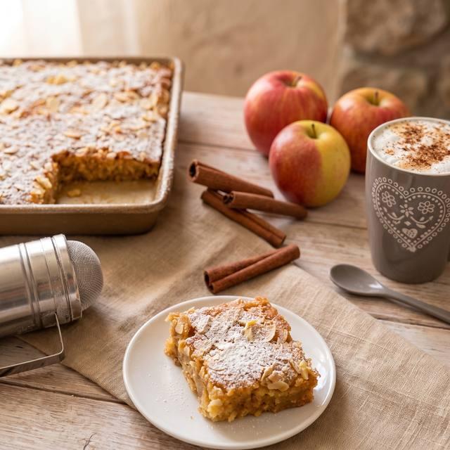 Vanille-Apfelkuchen vom Blech mit Puderzucker und Mandelblättchen bestreut, im Vordergrund ein Stück auf einem weißen Teller – Pampered Chef® Rezept